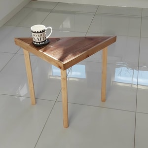 May include: A small, triangular wooden side table with a dark brown tabletop and light-coloured legs. A white mug with black text and a coaster sit on the table. The table is on a tiled floor.