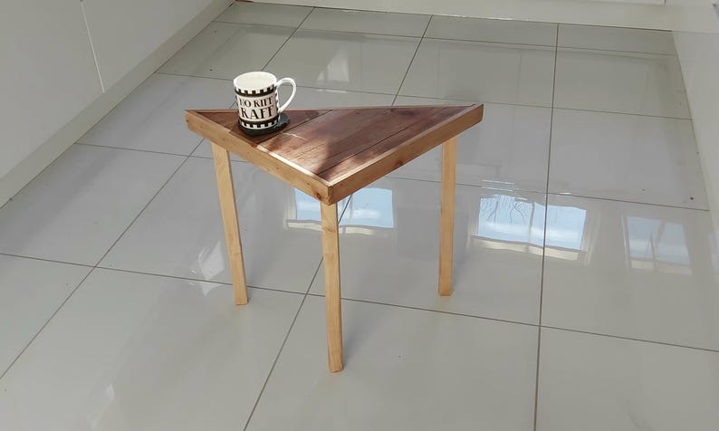 May include: A small, triangular wooden side table with a dark brown tabletop and light-coloured legs. A white mug with black text and a coaster sit on the table. The table is on a tiled floor.