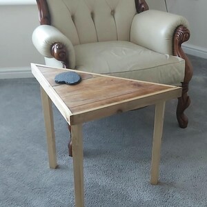 May include: A small, triangular wooden side table with a natural wood finish. The table has four light-coloured legs and a dark coaster on top. The table is approximately 61 cm long and 61 cm wide.