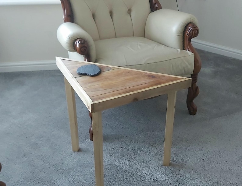 May include: A small, triangular wooden side table with a natural wood finish. The table has four light-coloured legs and a dark coaster on top. The table is approximately 61 cm long and 61 cm wide.