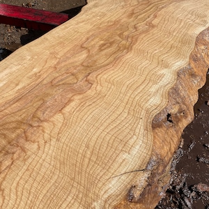 May include: A light-colored, unfinished wood slab with a natural edge. The wood grain is visible, with wavy lines and variations in color. The slab appears to be freshly cut and ready for use in woodworking projects.