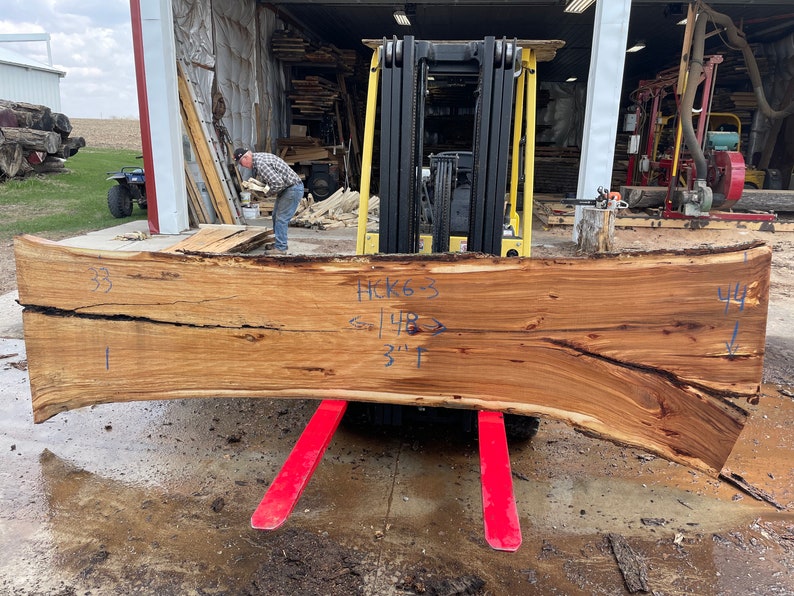 K&ouml;nnte beinhalten: Ein gro&szlig;es, gebogenes St&uuml;ck Holz mit nat&uuml;rlicher Oberfl&auml;che. Das Holz hat einen Riss, der durch die Mitte verl&auml;uft, und ist mit den Zahlen 33, 44 und dem Text "HCK63 < 14B > 3"1" gekennzeichnet. Das Holz wird von einem Gabelstapler angehoben.