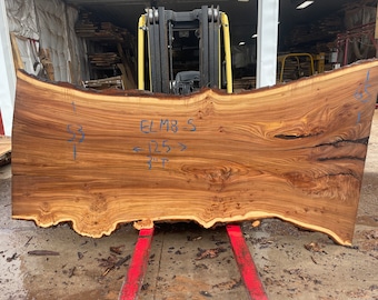 May include: A large slab of wood with a live edge, measuring 125 inches long and 3 inches thick. The wood is a light brown color with a natural grain pattern. The wood is labeled "ELM 8-S".