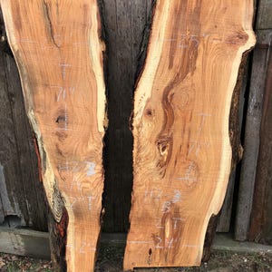 Pu&ograve; includere: Due tavole di legno non rifinite con bordi naturali e venature naturali. Le tavole sono marrone chiaro con bordi marroni e neri pi&ugrave; scuri. Segni bianchi indicano le misure.