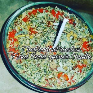 May include: A clear glass candle filled with a mix of dried herbs, red granules, and glitter. A white candle wick is centered in the jar. The text "The Voodoo Priestess Filed Road opener Candle" is written on the candle.