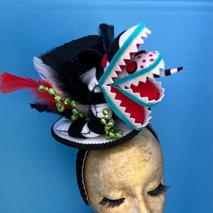 May include: A black and white top hat with a red and white felt monster with a blue mouth on top. The hat is decorated with green and red feathers and beads.