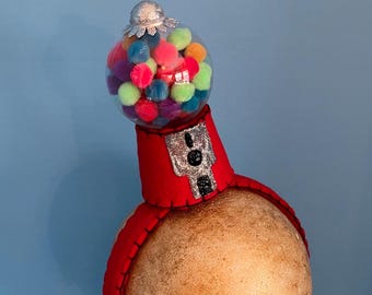 May include: A whimsical red headband with a silver and black accent, topped with a clear globe filled with colorful pom-poms. The headband is designed to resemble a gumball machine, creating a playful and unique accessory.