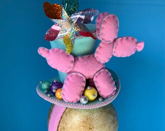 May include: A pink felt poodle with a blue felt top hat. The hat is decorated with a colorful pinwheel and silver and gold ornaments.