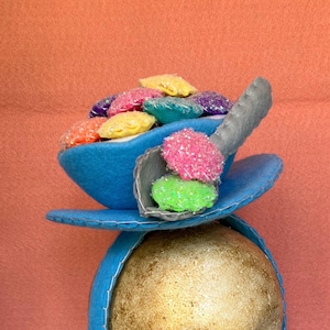 May include: A blue felt hat shaped like a teacup with a spoon and colorful felt cereal pieces on top.