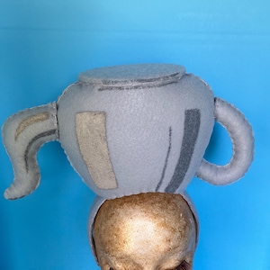 May include: A whimsical light gray felt teapot hat with a curved handle and spout, and a lid. The hat is adorned with lighter gray and darker gray felt accents. The hat is displayed on a mannequin head with long eyelashes and red lipstick.