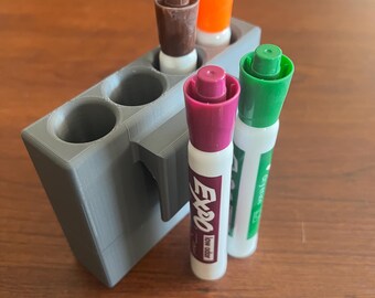 May include: A grey plastic marker holder with four slots for markers. The holder has a hook on the side. There are four markers in the holder: brown, orange, purple, and green.
