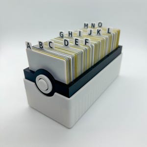 May include: A white and black rotary card file with alphabetized index tabs. The file contains a stack of cards with yellow and gray edges. The file has a rotating wheel on the side for easy access to the cards.