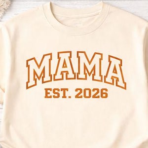 May include: Cream-colored t-shirt with the word "MAMA" in a brown, collegiate-style font, with "EST. 2026" below. The shirt is displayed with jeans, a candle, a gift box, and flowers.