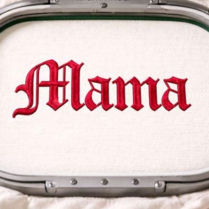 May include: An embroidery hoop with the word "Mama" stitched in red, gothic-style lettering. The fabric is white and stretched taut within the hoop. A spool of red thread is visible.