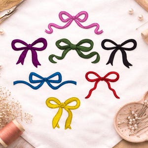 May include: Embroidered bow designs in various colors, including purple, green, black, blue, red, and yellow, are displayed on a white fabric. The bows are arranged in a decorative pattern, showcasing their different shapes and styles. A spool of thread and sewing notions are in the background.
