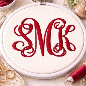 Op de afbeelding: Een geborduurd monogram in een witte borduurring. Het monogram toont de letters "SMK" in levendig rood, met een decoratief, vloeiend schrift. De stof is lichtbeige en de ring is wit. De afbeelding suggereert een knutselproject.