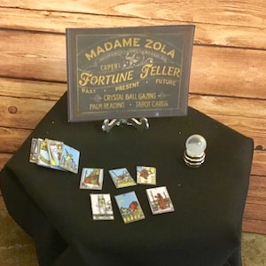 May include: A black sign with gold lettering that reads "Madame Zola Expert Fortune Teller Past Present Future Crystal Ball Gazing Palm Reading Tarot Cards" is on a stand with a crystal ball and tarot cards on a black cloth surface.