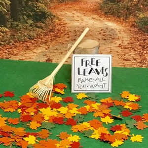 May include: A miniature wooden rake rests near a sign that reads "FREE LEAVES RAKE ALL YOU WANT." Colorful paper leaves in shades of red, orange, yellow, and green are scattered on a green surface, evoking an autumn theme.