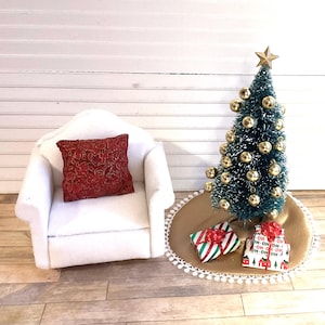 May include: A miniature white armchair with a red patterned pillow sits next to a small Christmas tree decorated with gold ornaments. The tree is on a gold and white tree skirt with wrapped presents underneath.
