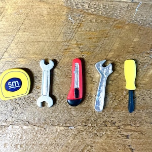 May include: A collection of miniature tools, including a yellow tape measure with "5m" printed on it, two silver wrenches, a red utility knife, and a yellow screwdriver. The tools are arranged on a wooden surface.