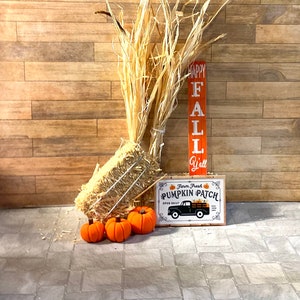 May include: A miniature hay bale with three orange pumpkins in front of a wooden wall. A sign reads "Happy Fall Y'all" and another sign reads "Farm Fresh Pumpkin Patch Open Daily".