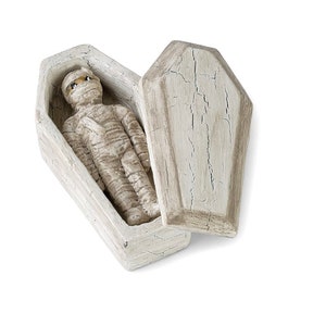 May include: A white wooden coffin with a mummy figurine inside. The coffin is open and the lid is lying next to it.