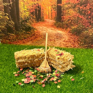 May include: A miniature autumn scene featuring two hay bales, a small wooden rake, and scattered colorful leaves on a green surface. The background shows a path through a forest with fall foliage.