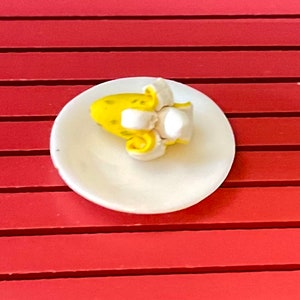 May include: A small white plate with a yellow banana and white frosting on top. The plate is on a red surface.