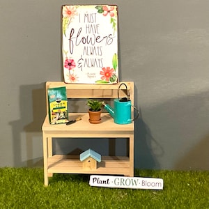 May include: A miniature wooden potting bench with a sign that reads "I MUST HAVE FLOWERS ALWAYS & ALWAYS." The bench holds a bag of garden soil, a small potted plant, a blue watering can, and a birdhouse. The sign also has the text "-CLAUDE MONET."