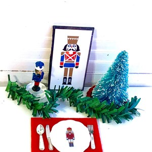 May include: A red felt placemat with a white plate featuring a nutcracker design. A silver fork and knife are placed on the placemat. A green garland and a small blue Christmas tree are in the background.