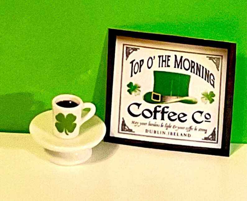 May include: A white coffee cup with a green shamrock design sits on a white saucer. Behind the cup is a framed sign that reads "Top O' The Morning Coffee Co. May your worries be light & your coffee be strong. Dublin, Ireland."