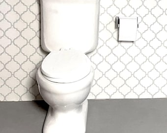 May include: White toilet with a closed lid and a white toilet paper holder with a roll of toilet paper on a white and gray patterned tile wall.