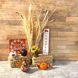 May include: A Thanksgiving-themed display with two bales of hay, a sign that reads "Happy Thanks Giving", a sign that reads "In all things give thanks", a small turkey figurine, a small pumpkin, and a bouquet of dried flowers.