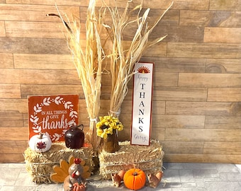 May include: A Thanksgiving-themed display with two bales of hay, a sign that reads "Happy Thanks Giving", a sign that reads "In all things give thanks", a small turkey figurine, a small pumpkin, and a bouquet of dried flowers.