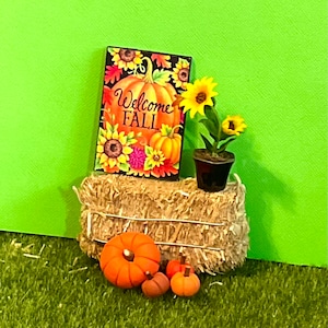 May include: A miniature hay bale with a welcome fall sign, a potted sunflower plant, and three small pumpkins. The sign has a pumpkin and sunflowers on it.