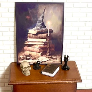 May include: A framed painting of a stack of old books with a witch's hat on top. The books are on a table with a skull, a small witch's hat, a book, and a candle holder.