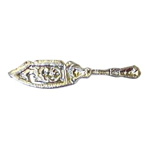 May include: A small, silver-toned metal charm shaped like a cake server or spatula. The flat, triangular blade features an embossed, intricate scrollwork pattern, while the handle is slender with a textured, decorative finish and a flared end.