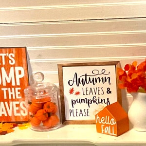 May include: A white shelf with fall decorations. There are two signs, one that says "Let's Jump in the Leaves" and another that says "Autumn Leaves & Pumpkins Please". There is a glass jar filled with small orange pumpkins, a white vase with orange leaves, and a small orange house sign that says "Hello Fall".
