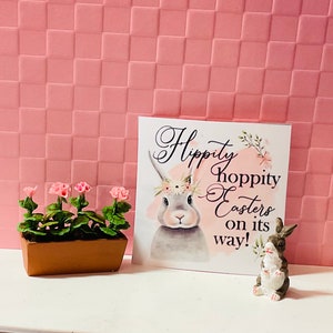 May include: A white card with the text "Hippity Hoppity Easter on its way!" and a gray bunny rabbit illustration. A small brown planter with four pink flowers sits in front of the card.
