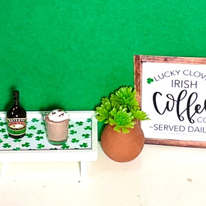 May include: A miniature white tray with a green shamrock pattern, holding a bottle of Baileys Irish Cream and a glass of Irish coffee with whipped cream and chocolate sprinkles. A small wooden sign with the text "Lucky Clover Irish Coffee Co. -Served Daily- " is on the right side of the image. A small green succulent plant in a terracotta pot is in the center of the image.