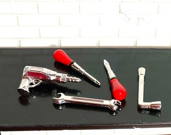 May include: A set of four silver tools on a black surface. The tools include a drill, a wrench, a screwdriver, and a socket wrench. The screwdriver and drill have red handles.