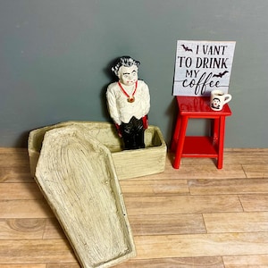 May include: A miniature coffin with a lid, a vampire figurine standing in the coffin, and a sign that reads "I vant to drink my coffee" with bats on either side of the text. The sign is on a red stool with a miniature coffee cup.