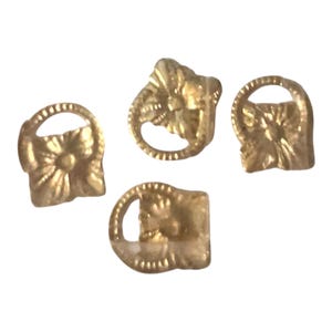 May include: Four small gold-toned metal charms, each featuring a square base with a raised floral design and a circular loop at the top. The metallic surface has a reflective finish, and the items are arranged in a cluster against a plain background.