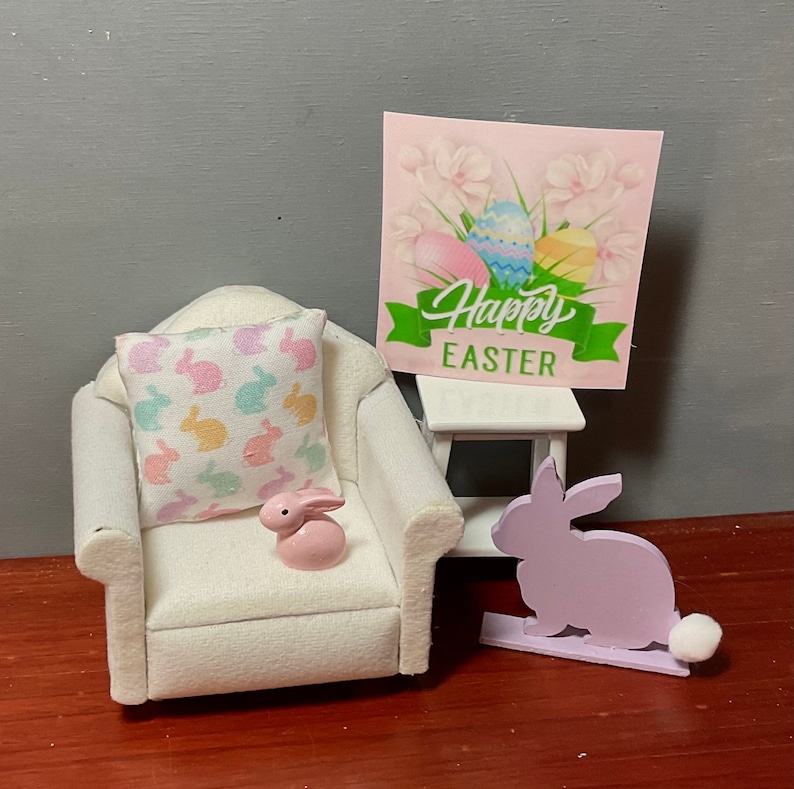 May include: A white armchair with a pillow featuring a pastel bunny pattern. A pink bunny figurine sits on the chair. A small white table with a sign that reads "Happy Easter" and a purple bunny figurine are also in the image.