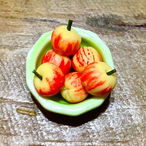 May include: A miniature light green bowl containing six small, round, yellow and red striped apples. Each apple features a tiny dark green stem protruding from the top. The fruit is arranged in a cluster within the bowl, displaying a smooth texture.