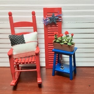 May include: A miniature patriotic scene featuring a red rocking chair with a flag-themed pillow, a red shutter with a star, and a blue table with a flower box. The scene is set on a red surface.