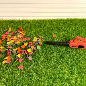 May include: A red and black toy leaf blower blowing a pile of colorful fall leaves on a green artificial grass surface.