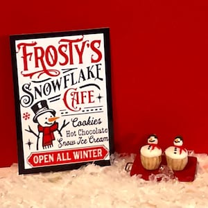May include: A sign for Frosty's Snowflake Cafe with a snowman illustration, offering cookies, hot chocolate, and snow ice cream. The sign reads "Open All Winter." Two snowman cupcakes sit on a red sled.