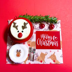 May include: A miniature Christmas table setting with a reindeer cake, a plate with a reindeer design, and a red card that says "Merry Christmas" with a heart.