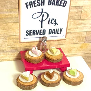 May include: A wooden sign with the text "Fresh Baked Pies Served Daily" is displayed above a table with six miniature pies on wooden rounds. The pies are topped with various fillings, including chocolate, fruit, and cream.